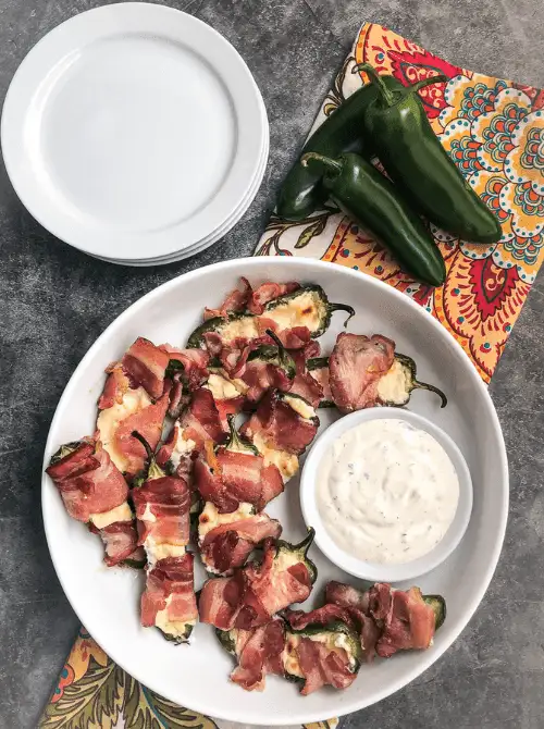 Bacon-wrapped jalapeño poppers served with ranch dip in a white bowl—spicy, crowd-pleasing tailgate food on a budget perfect for game day gatherings.