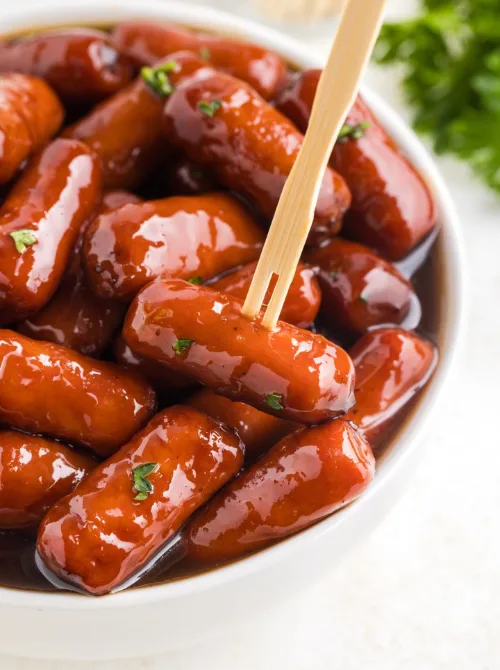 Slow-cooked little smokies coated in a glossy grape jelly and BBQ sauce glaze, served in a white bowl—an easy and nostalgic favorite for football party snacks on a budget.