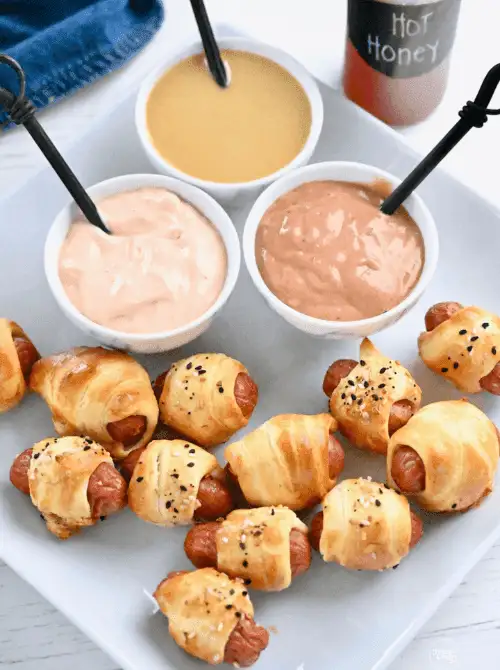 Mini pigs in a blanket topped with seasoning, served with three dipping sauces—easy tailgate food on a budget that's perfect for sharing with a crowd.