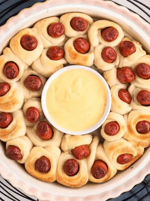 Golden pigs in a blanket arranged in a ring around a bowl of cheese dip—an easy and festive favorite for football party snacks on a budget.