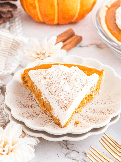 Slice of creamy pumpkin pie topped with whipped cream and cinnamon, served on a scalloped plate with fall decor—an easy and elegant solution for what to do with leftover pumpkin puree during the holidays.