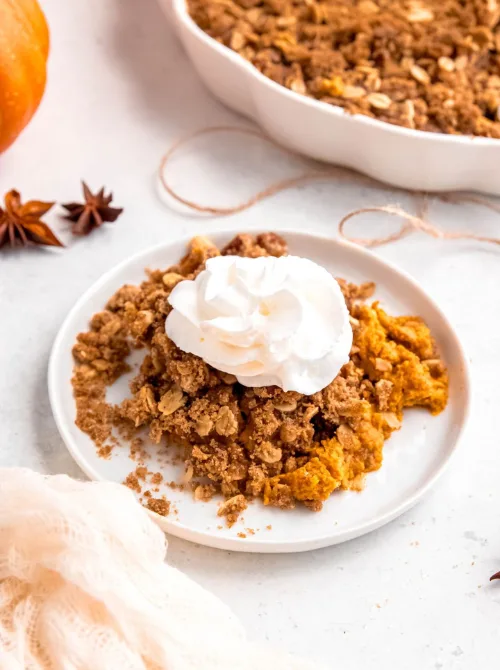 Plate of pumpkin crisp topped with whipped cream, featuring a golden oat crumble and spiced pumpkin filling—a cozy fall dessert and easy way to use leftover pumpkin puree.