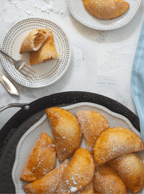 Golden-brown pumpkin empanadas dusted with powdered sugar on a serving tray and dessert plates—an easy idea for what to make with leftover pumpkin puree and a delicious twist on classic pumpkin desserts.