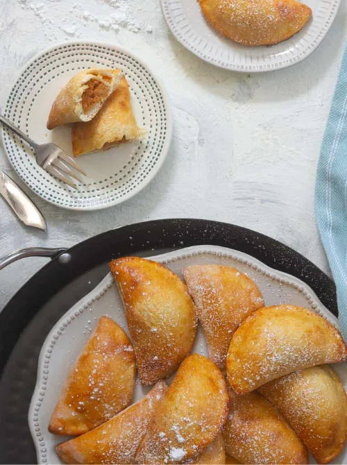 Golden-brown pumpkin empanadas dusted with powdered sugar on a serving tray and dessert plates—an easy idea for what to make with leftover pumpkin puree and a delicious twist on classic pumpkin desserts.