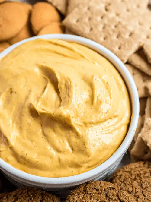Close-up of creamy pumpkin dip in a white bowl surrounded by graham crackers, gingersnaps, and vanilla wafers—an easy no-bake pumpkin dessert and fun way to use leftover pumpkin puree.