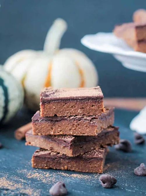 Stack of pumpkin fudge brownies with a rich chocolate topping, dusted with cinnamon and surrounded by fall decor—an indulgent vegan dessert and creative way to use leftover pumpkin puree.S