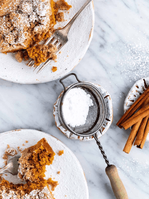 Pumpkin crumb cake dusted with powdered sugar on rustic plates with forks, cinnamon sticks, and a vintage sifter—perfect inspiration if you're wondering what to make with leftover pumpkin puree and want cozy pumpkin cakes and cupcakes.
