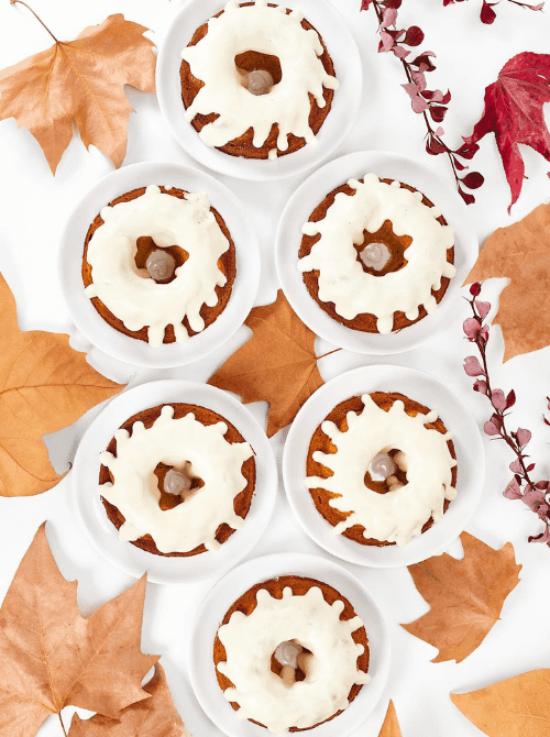 Mini pumpkin cakes topped with white icing and caramel centers, arranged on small plates with fall leaves—an adorable autumn dessert idea for what to make with leftover pumpkin puree in the form of pumpkin cakes and cupcakes.
