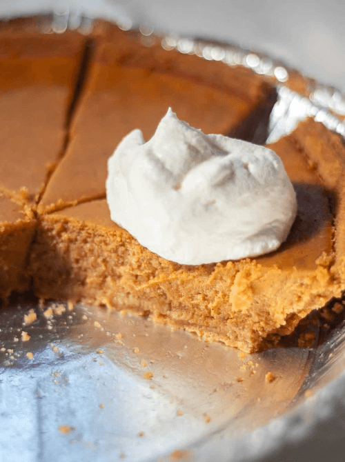 Close-up of a creamy pumpkin pie with a thick dollop of whipped cream on top, partially sliced in a foil pie tin—an inviting way to use leftover pumpkin puree in an easy homemade pumpkin pie.