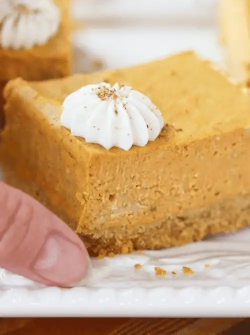 Thick slice of pumpkin cheesecake bar with graham cracker crust and whipped cream topping, being lifted from a serving plate — a rich and easy pumpkin cheesecake dessert made with leftover pumpkin purée.D