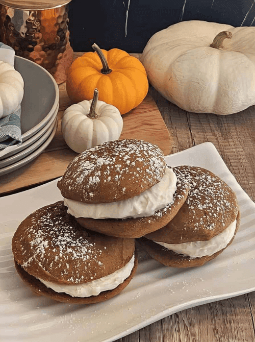 Three pumpkin whoopie pies filled with whipped cream and dusted with powdered sugar, served on a white platter with mini pumpkins in the background—a fun and festive way to use leftover pumpkin puree for fall treats.