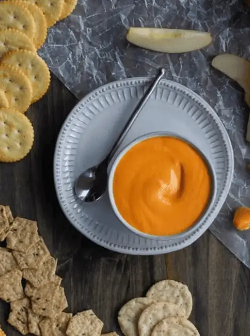 Bright orange roasted red pepper dip in a white bowl on a plate, surrounded by crackers, apple slices, and chips—perfect for easy game day dips that bring bold flavor and fun to the snack table.