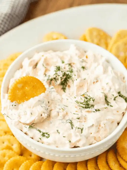 A creamy smoked salmon dip garnished with fresh dill and served with buttery round crackers, making it a crowd-pleasing choice for football party appetizers or easy snacks to feed a crowd.