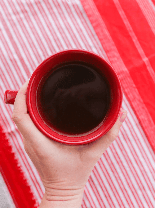 Hand holding a red mug filled with warm spiced apple cider, set against a festive red-striped background—simple, soothing, and a classic choice for easy fall recipes and nostalgic things to make with apples.
