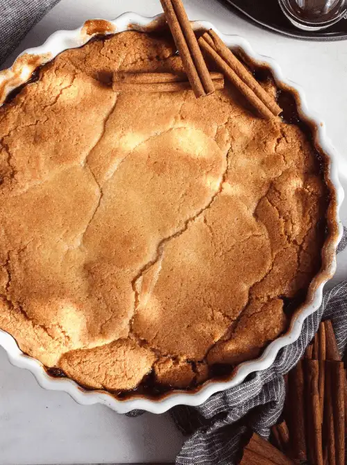 Golden-brown apple cobbler in a scalloped baking dish, topped with cinnamon sticks—an easy and comforting fall dessert and one of the coziest things to make with apples.