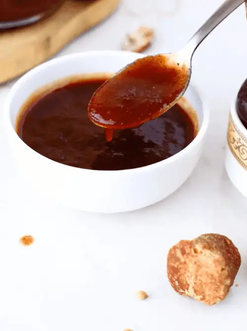 A rich, dark red tamarind sauce drizzled from a spoon into a white bowl, surrounded by raw tamarind pieces—perfect as a bold, tangy dip for game day snacks, football party appetizers, or a frugal twist on finger foods.