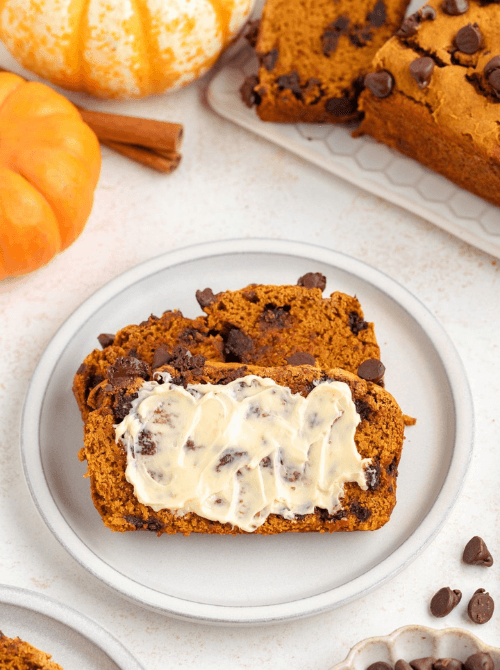 Two slices of chocolate chip pumpkin bread on a white plate, one topped with butter, surrounded by pumpkins and cinnamon sticks — perfect for ideas on what to do with leftover pumpkin puree and inspiration for easy pumpkin bread recipes.