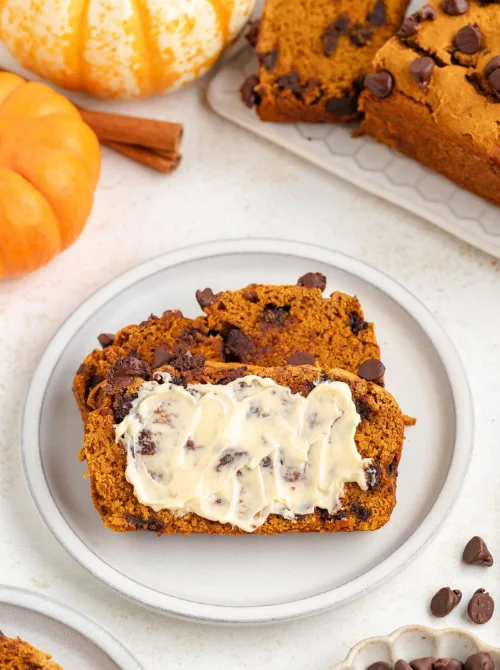 Two slices of chocolate chip pumpkin bread on a white plate, one topped with butter, surrounded by pumpkins and cinnamon sticks — perfect for ideas on what to do with leftover pumpkin puree and inspiration for easy pumpkin bread recipes.