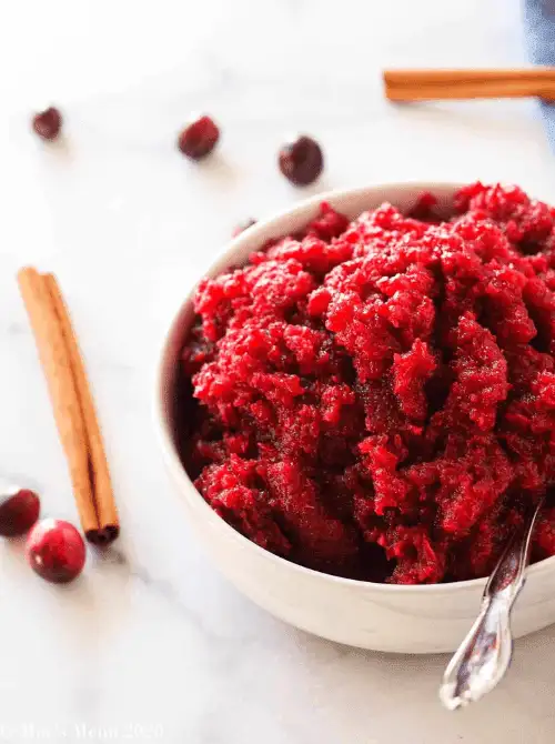 Bowl of apple cranberry sauce with a coarse texture, surrounded by cinnamon sticks and fresh cranberries, a festive budget-friendly Thanksgiving side.