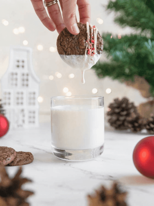 Hand dipping a decorated brownie cookie into a glass of milk, a festive DIY Christmas gift idea with cookies and bars.