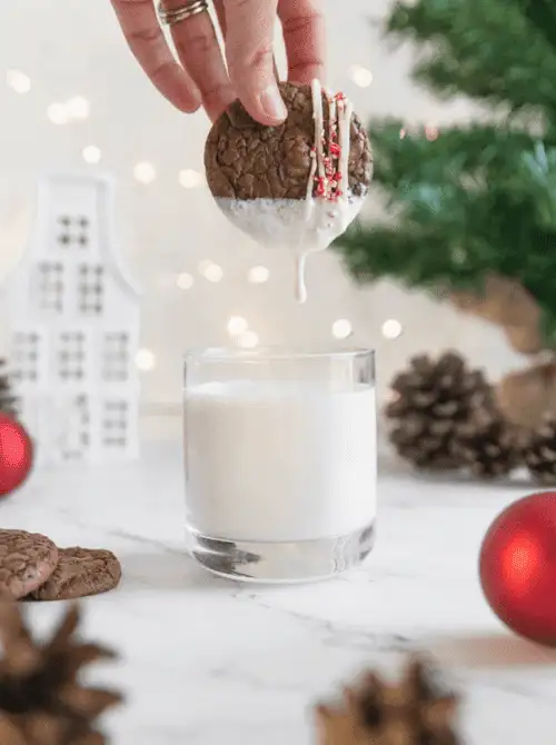 Hand dipping a decorated brownie cookie into a glass of milk, a festive DIY Christmas gift idea with cookies and bars.
