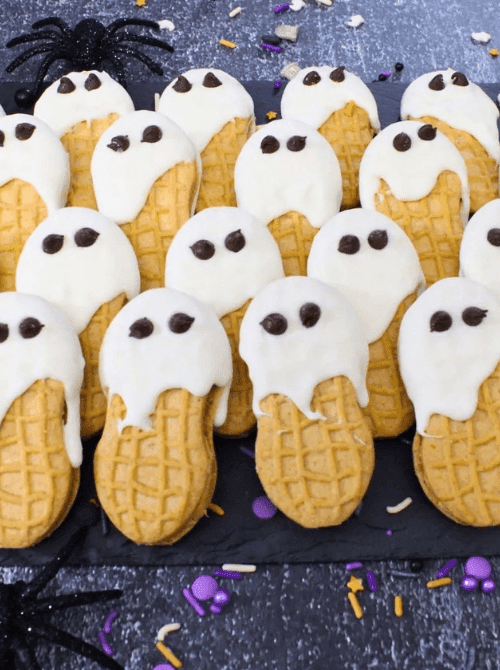 No-Bake Halloween Treats featuring Nutter Butter cookies dipped in white chocolate to resemble ghosts, decorated with chocolate chip eyes and arranged on a dark tray with festive Halloween sprinkles and spider props.