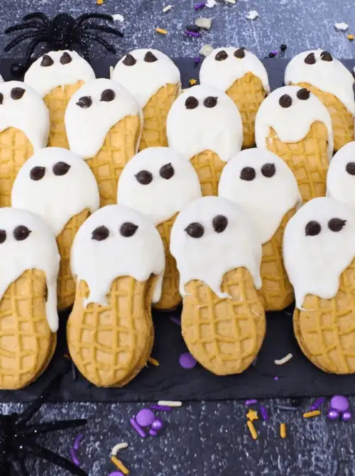 No-Bake Halloween Treats featuring Nutter Butter cookies dipped in white chocolate to resemble ghosts, decorated with chocolate chip eyes and arranged on a dark tray with festive Halloween sprinkles and spider props.