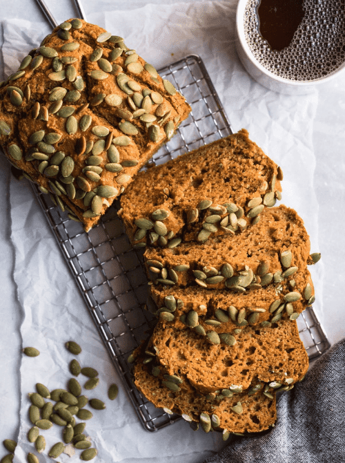 Sliced loaf of pumpkin bread topped with pumpkin seeds, displayed on a cooling rack beside a cup of coffee and scattered seeds.
