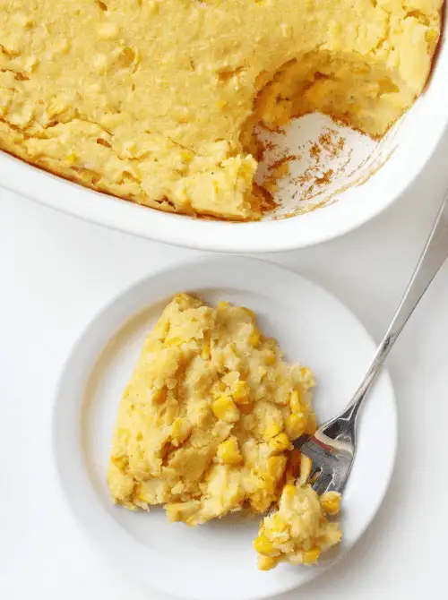 White baking dish and plate with a serving of creamy corn casserole made with whole kernels, a simple budget-friendly Thanksgiving side.