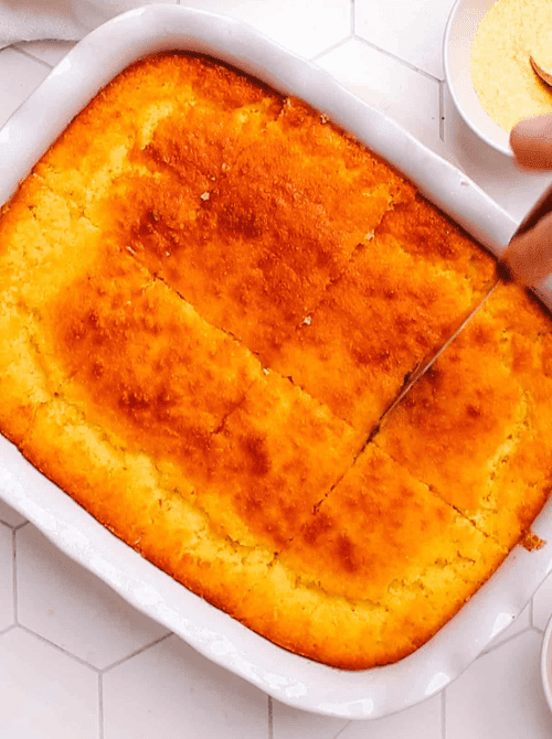 Golden baked cornbread casserole being sliced in a white dish, a warm and budget-friendly Thanksgiving side.
