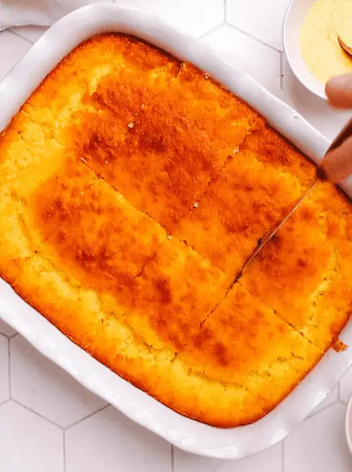 Golden baked cornbread casserole being sliced in a white dish, a warm and budget-friendly Thanksgiving side.