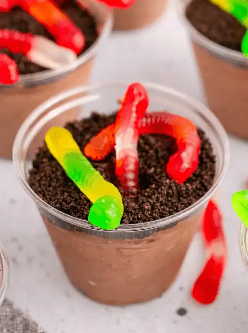 No-Bake Halloween Treats made with chocolate pudding layered under crushed Oreo “dirt,” topped with colorful gummy worms in a clear plastic cup for a spooky, kid-friendly dessert.