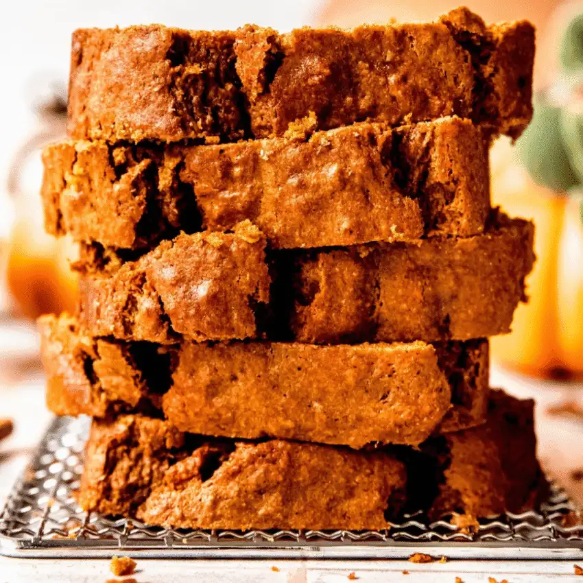 Stack of thick, moist pumpkin bread slices on a cooling rack, with a warm autumn background and blurred pumpkins.