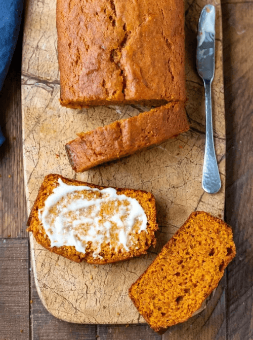 Freshly baked pumpkin bread loaf on a wooden cutting board with slices, one topped with melted butter, alongside a butter knife — a delicious example of what to do with leftover pumpkin puree and a classic choice for easy pumpkin bread recipes.