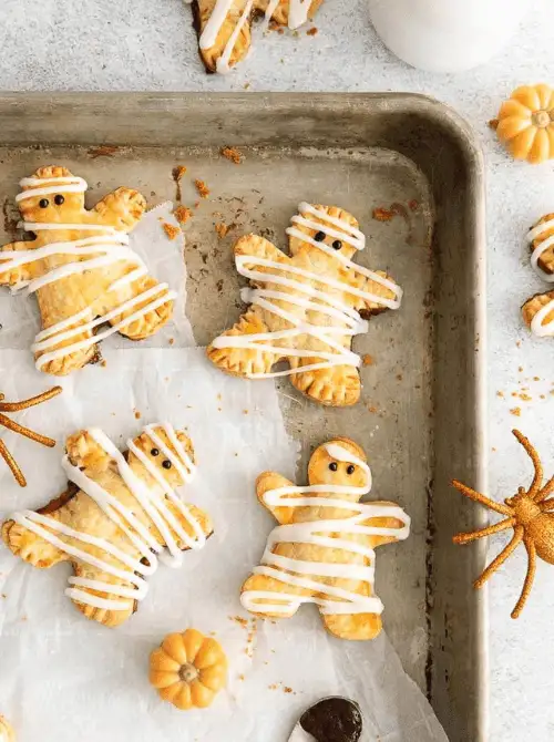 Mini mummy hand pies decorated with white icing and candy eyes, baked to golden perfection on a rustic tray, surrounded by mini pumpkins and Halloween props.