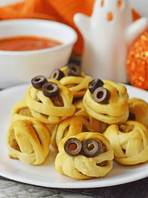 Mini meatballs wrapped in crescent dough to look like mummies, topped with sliced black olives for eyes—fun and kid-friendly make-ahead Halloween party food idea.