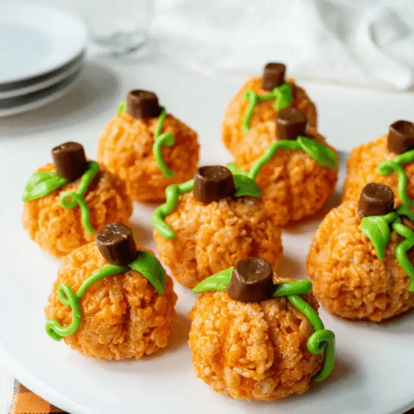 Pumpkin-shaped no-bake rice krispie treats topped with Rolos and green icing vines, arranged on a white plate—perfect for easy Halloween or fall party snacks.