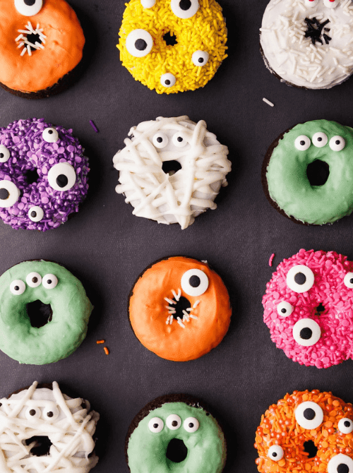 Colorful Halloween donuts decorated like monsters and mummies with bright frosting, candy eyes, and festive sprinkles, arranged in a neat grid on a dark background.