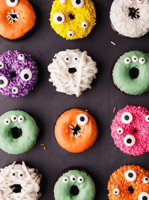 Colorful Halloween donuts decorated like monsters and mummies with bright frosting, candy eyes, and festive sprinkles, arranged in a neat grid on a dark background.