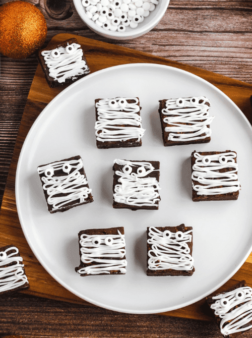 Brownie squares decorated with white icing “bandages” and candy eyes to look like mummies—easy and fun make-ahead Halloween party food perfect for kids and dessert tables.