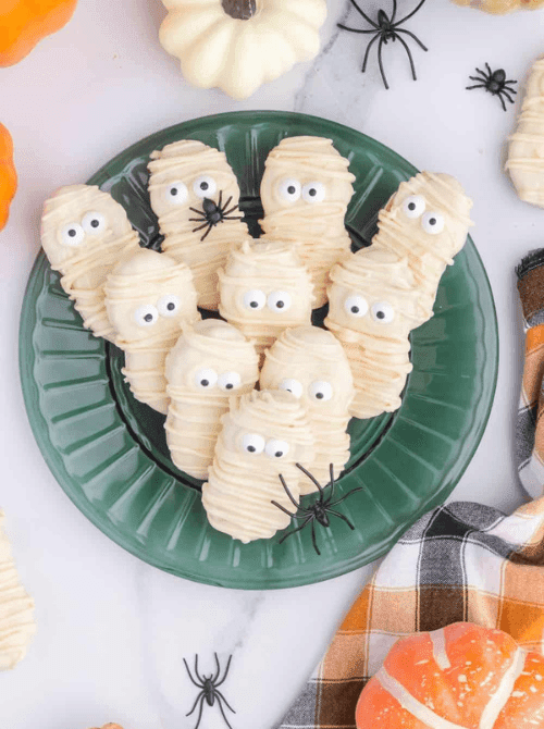 No-Bake Halloween Treats made with Nutter Butter cookies dipped in white chocolate and decorated like mummies with candy eyes, arranged on a green plate with festive plastic spiders.