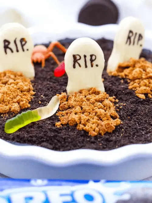 No-Bake Halloween Treats styled as a spooky graveyard dessert with crushed Oreo “dirt,” candy tombstones marked “RIP,” gummy worms, and cookie crumble mounds in a white pie dish.