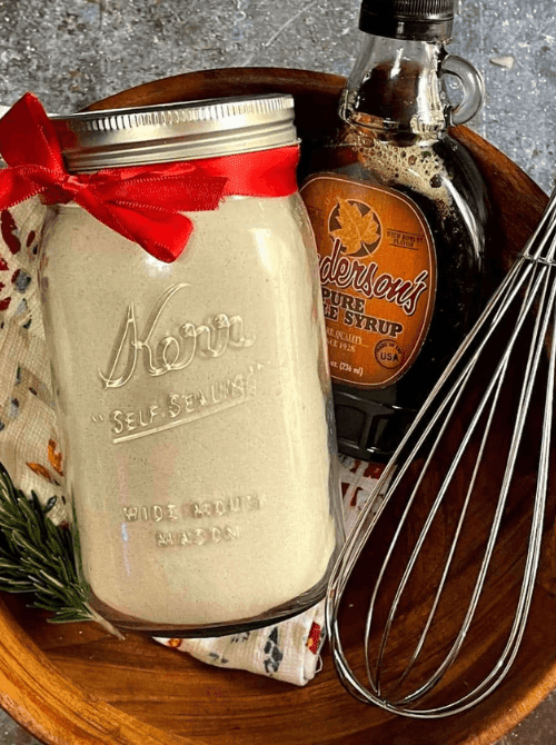 Homemade pancake mix in a mason jar tied with a red bow, displayed with maple syrup and a whisk, a cozy DIY Christmas gift and mason jar recipe.