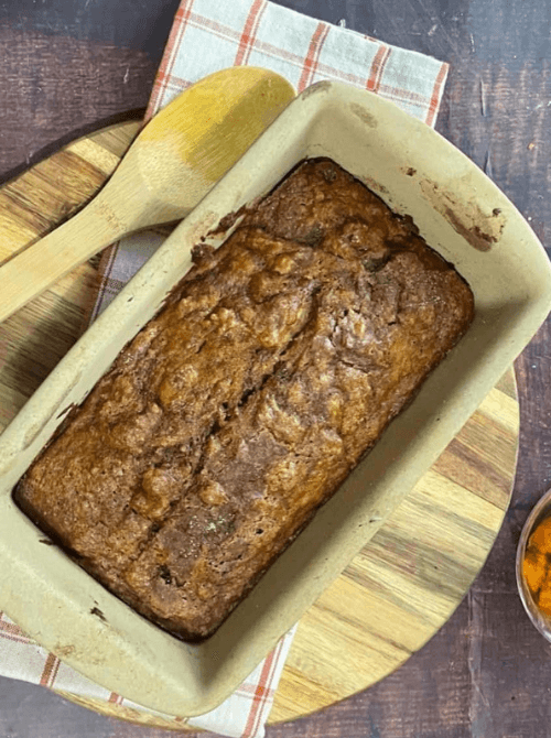 Freshly baked pumpkin bread loaf in a ceramic pan on a wooden board with a wooden spoon — a cozy fall bake and a tasty example of what to do with leftover pumpkin puree for easy pumpkin bread recipes.