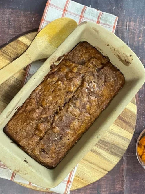 Freshly baked pumpkin bread loaf in a ceramic pan on a wooden board with a wooden spoon — a cozy fall bake and a tasty example of what to do with leftover pumpkin puree for easy pumpkin bread recipes.