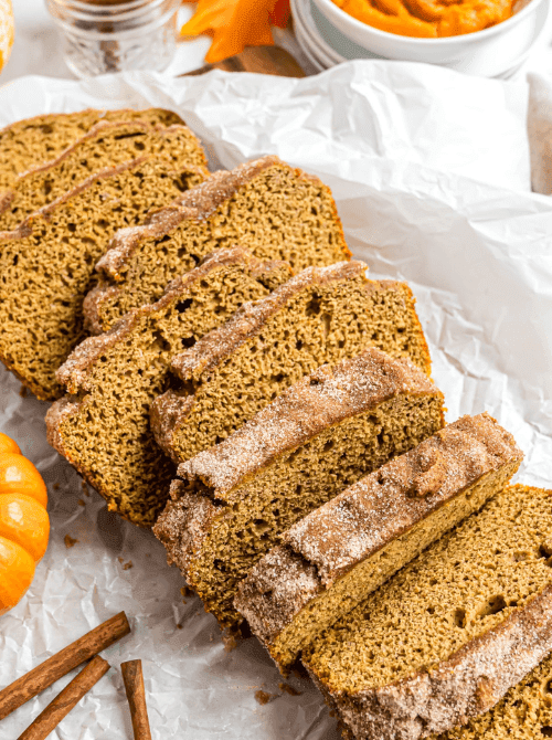 Several slices of cinnamon sugar-topped pumpkin bread arranged on parchment paper with mini pumpkins and cinnamon sticks — a sweet and spiced idea for what to do with leftover pumpkin puree and a cozy option for easy pumpkin bread recipes.