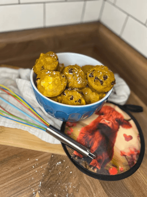 Blue bowl filled with bite-sized pumpkin chocolate chip muffins, dusted with powdered sugar, on a wooden counter with a whisk and cow-themed pot holder — a fun and tasty idea for what to do with leftover pumpkin puree and a creative twist on easy pumpkin bread recipes.