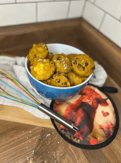 Blue bowl filled with bite-sized pumpkin chocolate chip muffins, dusted with powdered sugar, on a wooden counter with a whisk and cow-themed pot holder — a fun and tasty idea for what to do with leftover pumpkin puree and a creative twist on easy pumpkin bread recipes.