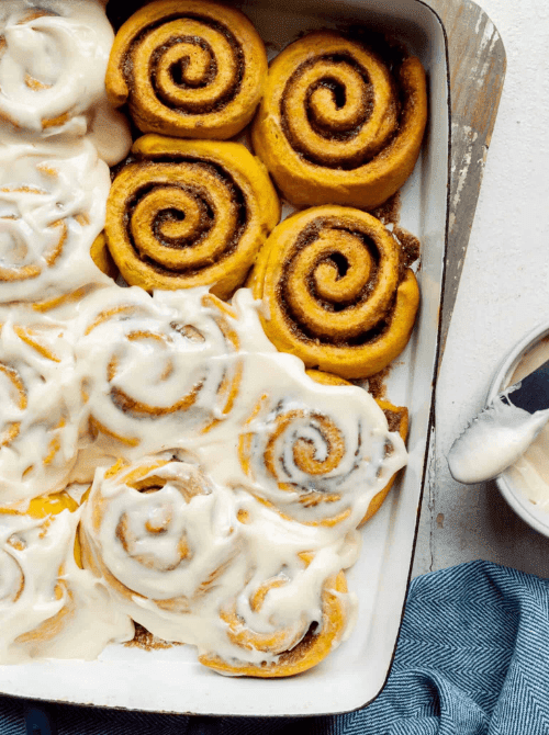 "Freshly baked pumpkin cinnamon rolls in a baking dish, half generously covered with creamy icing and half showing the warm, golden swirls of cinnamon-sugar filling — a cozy fall treat perfect for using leftover pumpkin puree.