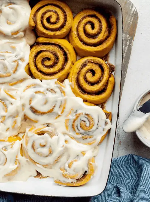 "Freshly baked pumpkin cinnamon rolls in a baking dish, half generously covered with creamy icing and half showing the warm, golden swirls of cinnamon-sugar filling — a cozy fall treat perfect for using leftover pumpkin puree.
